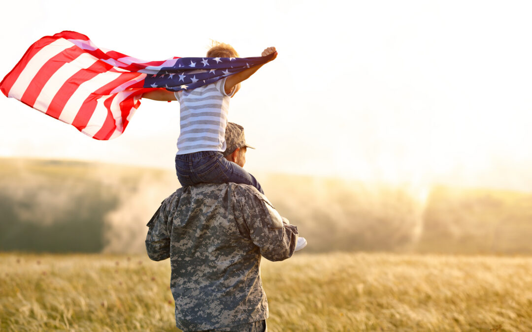 Veteran in military uniform carrying a child on their shoulders while the child waves an American flag in an open field at sunset, symbolizing family support and opportunities for veterans transitioning to civilian life and business ownership.